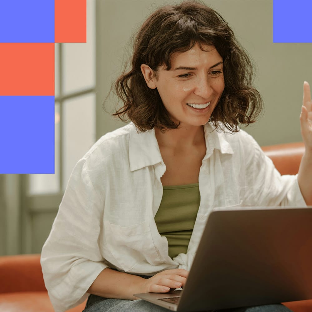 A smiling woman waves at her laptop screen during a video call, seated on an orange sofa with blue and red geometric shapes in the background.