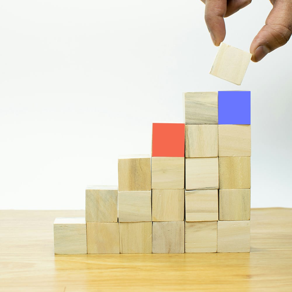 Hand placing a wooden block on a staircase-like stack of cubes, symbolizing growth or progress.
